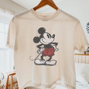 以下が含まれることがあります： アイボリー色のTシャツ。ミッキーマウスのイラストが黒、赤、白で描かれています。半袖のシャツは木製のハンガーに掛けられています。「Ivory」の文字が右下に印刷されています。