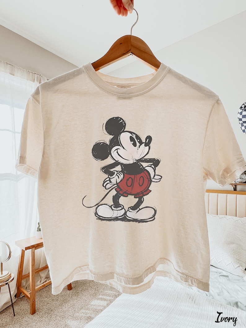 以下が含まれることがあります： アイボリー色のTシャツ。ミッキーマウスのイラストが黒、赤、白で描かれています。半袖のシャツは木製のハンガーに掛けられています。「Ivory」の文字が右下に印刷されています。