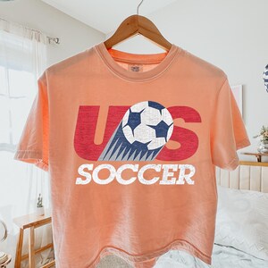 May include: A peach-colored short-sleeve t-shirt featuring a graphic print with the red letters U and S, a white and blue soccer ball replacing the letter S, and the word SOCCER in white block letters below the graphic on the front.