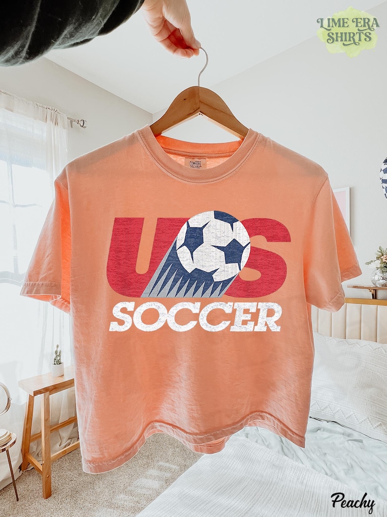 May include: A peach-colored short-sleeve t-shirt featuring a graphic print with the red letters U and S, a white and blue soccer ball replacing the letter S, and the word SOCCER in white block letters below the graphic on the front.
