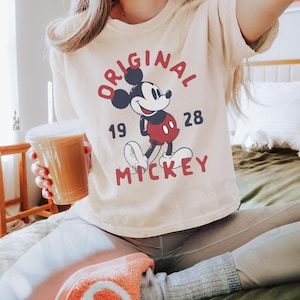 May include: Ivory-colored t-shirt featuring a graphic of Mickey Mouse with the text "ORIGINAL MICKEY 1928". The shirt has short sleeves and a crew neck. The image also includes a cup of coffee.