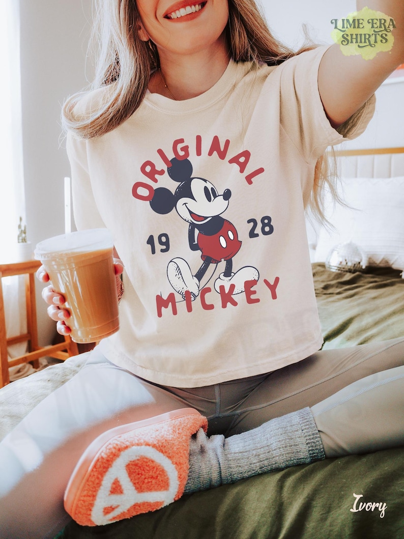 May include: Ivory-colored t-shirt featuring a graphic of Mickey Mouse with the text "ORIGINAL MICKEY 1928". The shirt has short sleeves and a crew neck. The image also includes a cup of coffee.