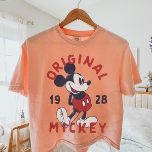 May include: Peach-colored t-shirt featuring a graphic of Mickey Mouse with the words "ORIGINAL 1928 MICKEY" in red. The shirt is hanging on a wooden hanger. The text "LIME ERA SHIRTS" is in the upper right corner.
