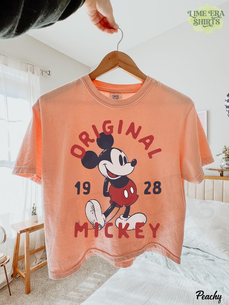 May include: Peach-colored t-shirt featuring a graphic of Mickey Mouse with the words "ORIGINAL 1928 MICKEY" in red. The shirt is hanging on a wooden hanger. The text "LIME ERA SHIRTS" is in the upper right corner.