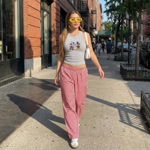 May include: A woman walking down a city sidewalk wearing a gray tank top with a Mickey Mouse graphic, red and white checkered pants, yellow sunglasses, and white sneakers. She carries a white purse.
