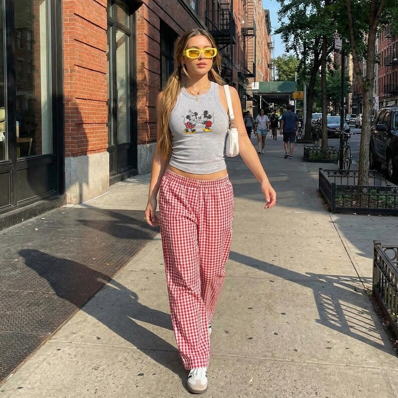 May include: A woman walking down a city sidewalk wearing a gray tank top with a Mickey Mouse graphic, red and white checkered pants, yellow sunglasses, and white sneakers. She carries a white purse.