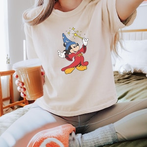 May include: Ivory-colored t-shirt featuring a cartoon Mickey Mouse dressed as a wizard, holding a wand with stars. The shirt is a neutral color. The word "Ivory" is printed on the bottom right corner of the image.