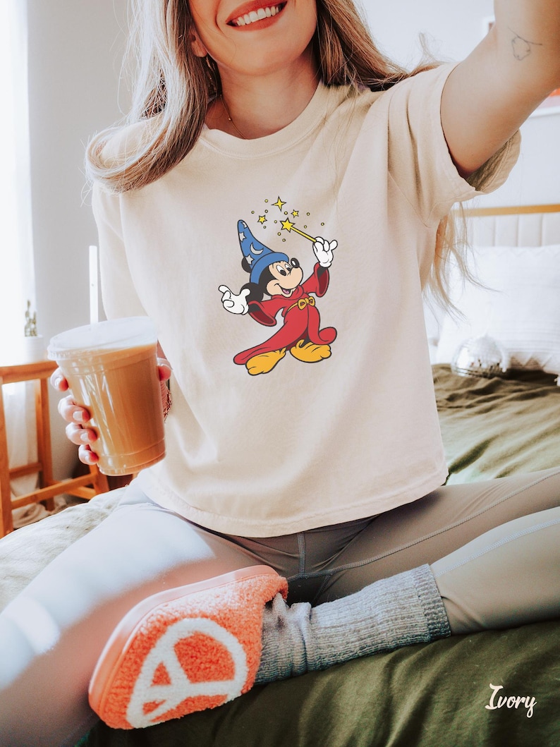 May include: Ivory-colored t-shirt featuring a cartoon Mickey Mouse dressed as a wizard, holding a wand with stars. The shirt is a neutral color. The word "Ivory" is printed on the bottom right corner of the image.