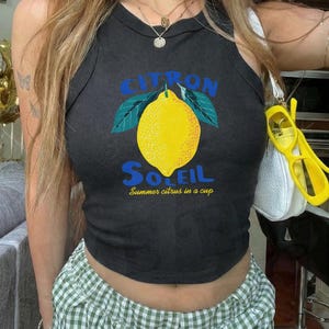 Puede incluir: Una camiseta de tirantes negra sin mangas con un gr&aacute;fico de un lim&oacute;n amarillo con hojas verdes. El texto dice CITRON SOLEIL y Summer citrus in a cup en letras azules y amarillas. La prenda tiene cuello redondo y un corte corto y ajustado.