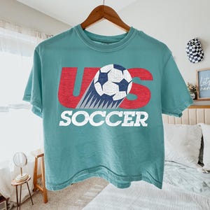 May include: A teal short-sleeve t-shirt featuring a graphic with the red letters U and S, a white and blue soccer ball replacing the letter O, and the word SOCCER printed in white block letters below the graphic on the front of the shirt.