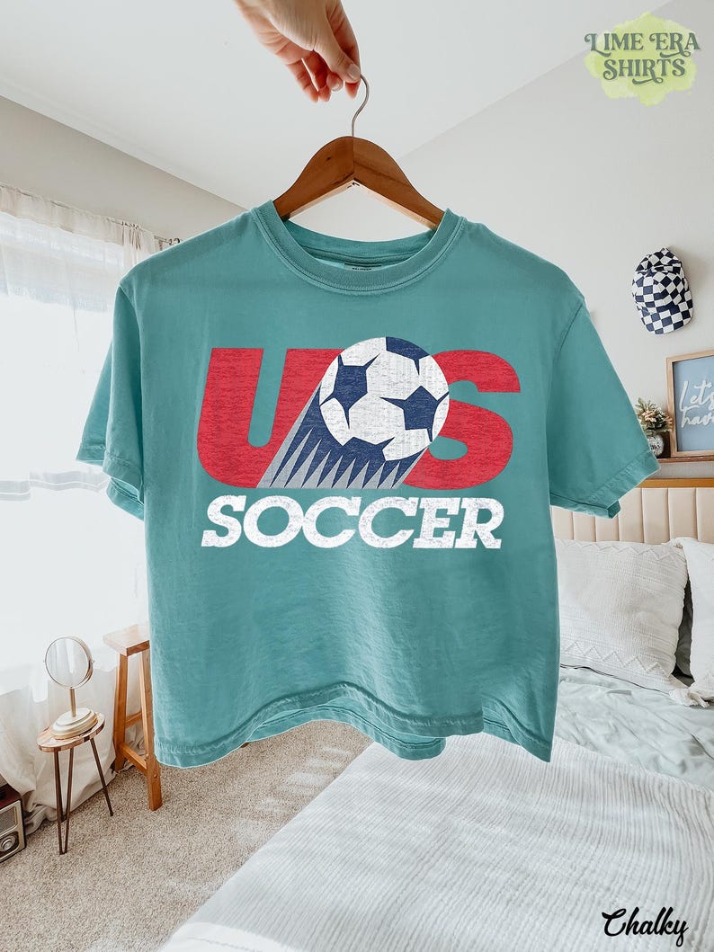 May include: A teal short-sleeve t-shirt featuring a graphic with the red letters U and S, a white and blue soccer ball replacing the letter O, and the word SOCCER printed in white block letters below the graphic on the front of the shirt.