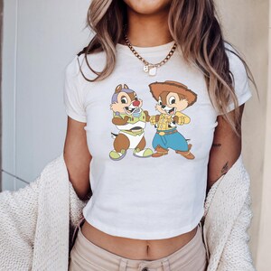 May include: White crop top featuring a cartoon graphic of two chipmunks, one dressed as a space ranger and the other as a cowboy. The chipmunks are in a playful pose. The shirt has a crew neck and short sleeves.