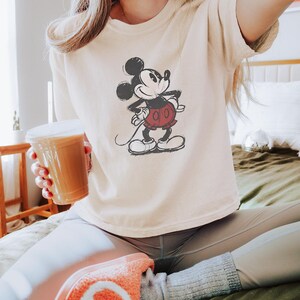 以下が含まれることがあります： アイボリー色のTシャツ。ミッキーマウスのイラストが黒、赤、白で描かれています。Tシャツはゆったりとしたフィット感で、半袖です。「Ivory」の文字が右下にプリントされています。