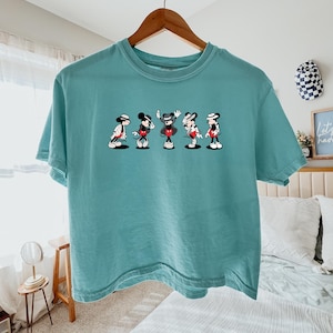 Puede incluir: Una camiseta de manga corta color verde azulado con un estampado gr&aacute;fico de cinco personajes de ratones de dibujos animados en varias poses, cada uno con sombrero y traje. La camiseta se muestra en una percha de madera.