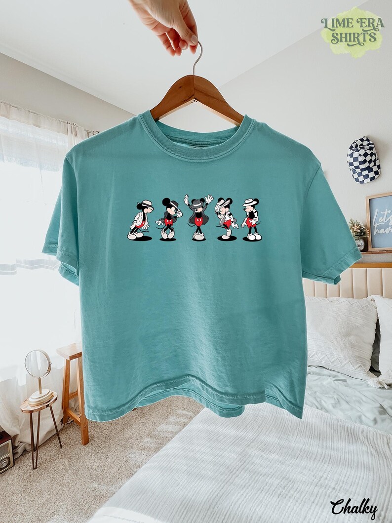 Puede incluir: Una camiseta de manga corta color verde azulado con un estampado gr&aacute;fico de cinco personajes de ratones de dibujos animados en varias poses, cada uno con sombrero y traje. La camiseta se muestra en una percha de madera.