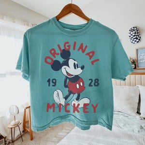 May include: Teal-colored t-shirt featuring a graphic of Mickey Mouse with the words "ORIGINAL MICKEY 1928" in red. The shirt is hanging on a wooden hanger. The image is taken indoors.