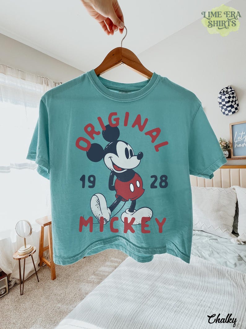 May include: Teal-colored t-shirt featuring a graphic of Mickey Mouse with the words "ORIGINAL MICKEY 1928" in red. The shirt is hanging on a wooden hanger. The image is taken indoors.