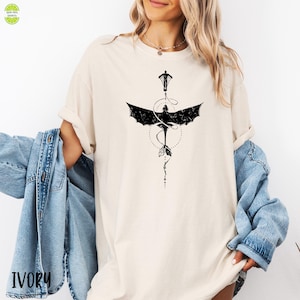 May include: Ivory t-shirt with a black dragon graphic, person, and dotted circle design. The word "IVORY" is printed at the bottom left. The shirt has short sleeves and a crew neck. The design features a dragon with outstretched wings. The shirt is a neutral colour.