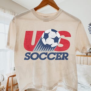 May include: An ivory short-sleeve t-shirt featuring a graphic print with the red letters U and S, a white and blue soccer ball, and the blue text SOCCER. The design has a distressed texture and is centered on the front of the garment.