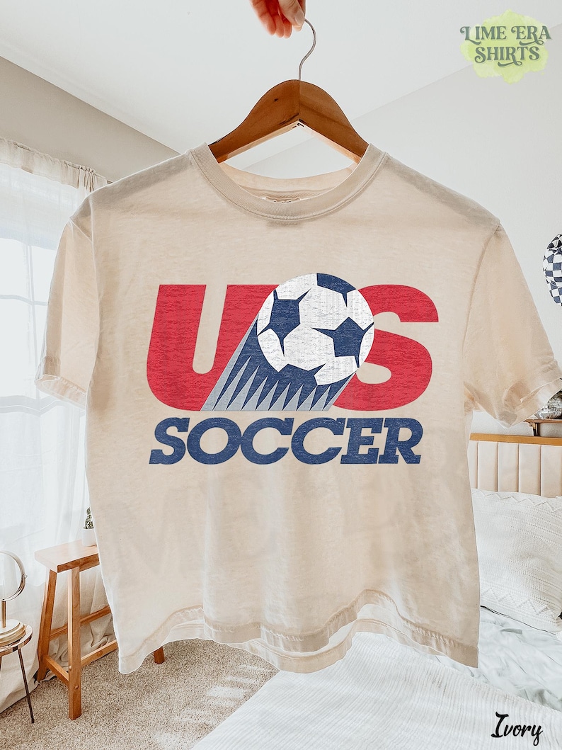 May include: An ivory short-sleeve t-shirt featuring a graphic print with the red letters U and S, a white and blue soccer ball, and the blue text SOCCER. The design has a distressed texture and is centered on the front of the garment.