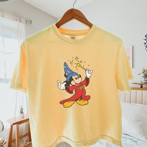 May include: Yellow t-shirt featuring a graphic of Mickey Mouse dressed as a sorcerer, holding a wand with stars. The shirt is hanging on a wooden hanger. The shirt is a light yellow color.