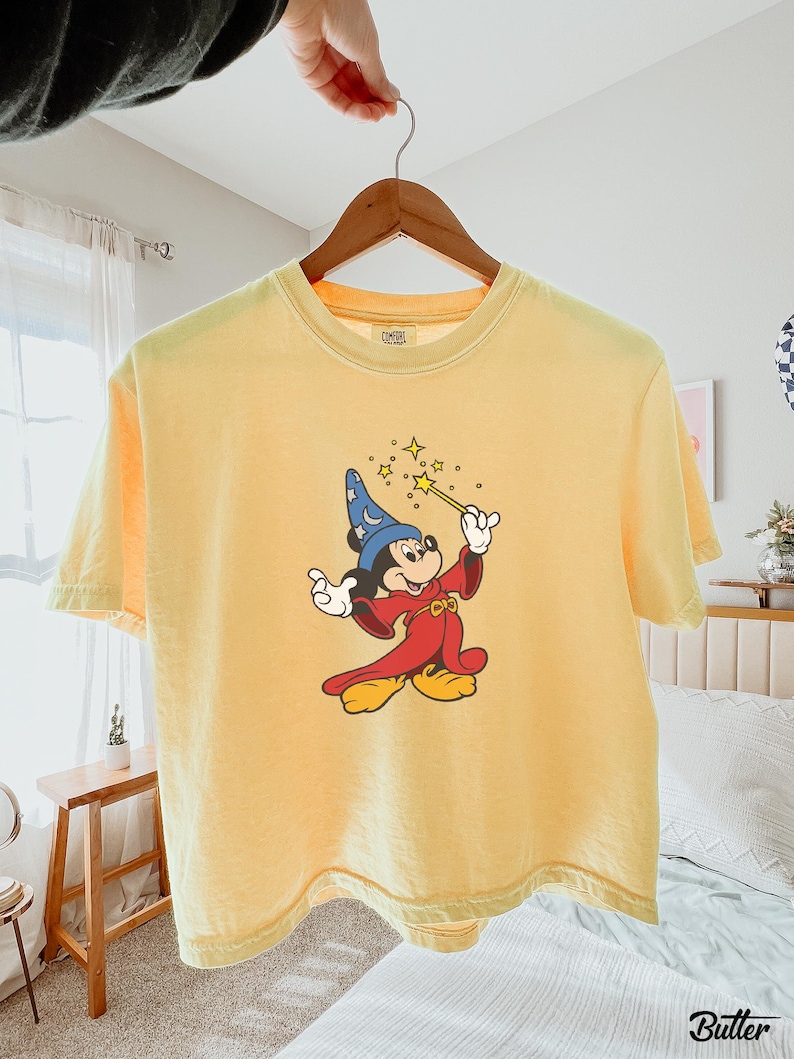 May include: Yellow t-shirt featuring a graphic of Mickey Mouse dressed as a sorcerer, holding a wand with stars. The shirt is hanging on a wooden hanger. The shirt is a light yellow color.