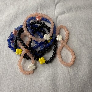 May include: A set of colorful beaded bracelets with flower charms. The bracelets are pink, blue, and black. The flower charms are white, yellow, and pink.