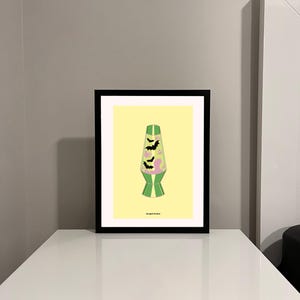 Puede incluir: Una impresión enmarcada de una lámpara de lava verde y rosa con murciélagos negros volando a su alrededor. El fondo es amarillo. El texto "Mogul Studio" está en la parte inferior de la impresión.