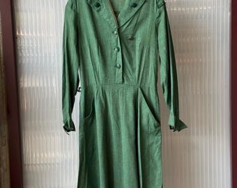 May include: A vintage green dress with long sleeves, a button-down front, and pockets. A matching green hat with a gold emblem is also present. The dress has a tailored fit and appears to be made of a textured fabric. The outfit is likely from a historical era.