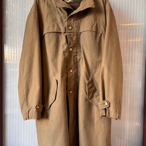 May include: A tan long-length coat featuring a button-front closure, a hood with a dark green and brown patterned lining, long sleeves with buttoned cuffs, and two front pockets. The coat is displayed on a wooden hanger.