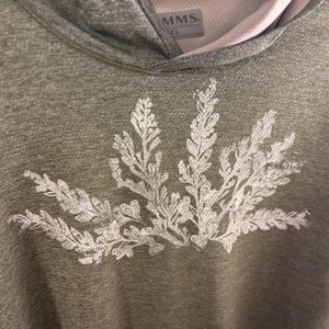 May include: Olive green hooded sweatshirt with a white fern design on the front. The sweatshirt has a light pink lining in the hood and a tag that reads "MMS XL". The fabric has a textured appearance.
