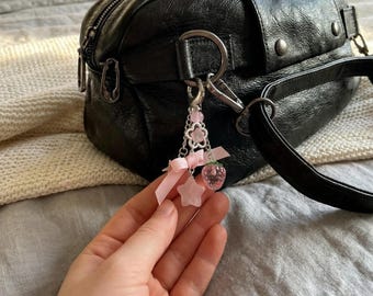 May include: A silver keychain charm with pink accents, including a flower, star, and strawberry. The charm is attached to a black leather bag. The charm has a pink ribbon and pink beads.