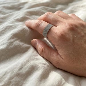 Könnte beinhalten: Ein silberner Ring mit gewebter Textur wird an einem Finger getragen. Der Ring hat ein breites Band und eine strukturierte Oberfläche. Der Ring besteht aus glänzendem Metall und wird auf einer Hand präsentiert.