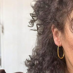 May include: A close-up of a person's ear wearing a gold-colored hoop earring. The earring has a teardrop shape. The person has dark, curly hair and is wearing a light green top.