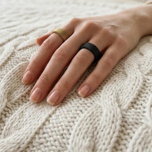 Op de afbeelding: Een hand met twee ringen: een goudkleurige ring en een zwarte ring. De ringen hebben een mesh-ontwerp. De hand rust op een crèmekleurige kabelgebreide stof.