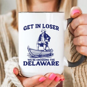 May include: White ceramic mug with a black handle and rim, featuring the text "GET IN LOSER" above an illustration of a man in a boat, with the text "WE'RE CROSSING THE DELAWARE" below. The mug is labeled "15oz".