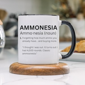May include: White ceramic mug with a black handle and rim, featuring the word "AMMONESIA" and a definition about forgetting how much ammo you have. The mug sits on a wooden coaster. The mug is 11oz.