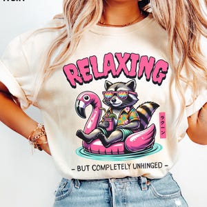 May include: Ivory-colored t-shirt featuring a raccoon wearing sunglasses and a Hawaiian shirt, relaxing on a pink flamingo float. The word "RELAXING" is above the raccoon, with "- BUT COMPLETELY UNHINGED -" below. Japanese text is on the right.