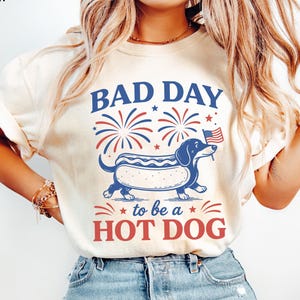 May include: Ivory-colored t-shirt with the text "BAD DAY to be a HOT DOG" in red and blue. The graphic features a dachshund hot dog with an American flag. Fireworks and stars complete the patriotic design.