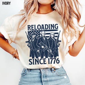 May include: Ivory-colored t-shirt featuring a navy blue graphic of three historical figures holding rifles, with an American flag background. The text "RELOADING SINCE 1776" is printed below the figures. The shirt is paired with light blue jeans.