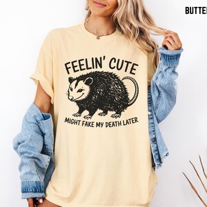 Może przedstawiać: Kremowa koszulka z czarną grafiką oposa i napisem "Feelin' Cute Might Fake My Death Later". Koszulka ma kr&oacute;tkie rękawy i luźny kr&oacute;j. Modelka ma na sobie dżinsową kurtkę.