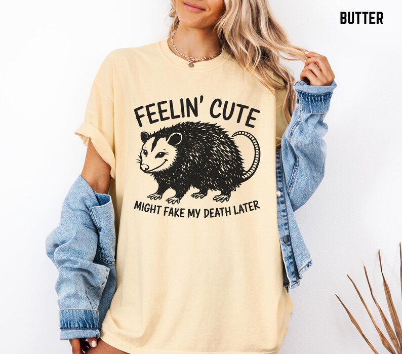 Może przedstawiać: Kremowa koszulka z czarną grafiką oposa i napisem "Feelin' Cute Might Fake My Death Later". Koszulka ma kr&oacute;tkie rękawy i luźny kr&oacute;j. Modelka ma na sobie dżinsową kurtkę.