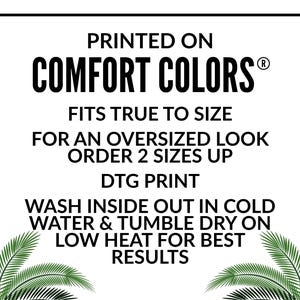 May include: White graphic with black text that reads "PRINTED ON COMFORT COLORS&reg;" and care instructions. The text advises to wash inside out in cold water and tumble dry on low heat. Palm fronds frame the text.