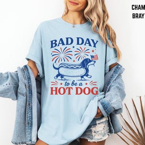 May include: Light blue t-shirt with a graphic of a dachshund hot dog, fireworks, and the text "BAD DAY to be a HOT DOG" in red and blue. The shirt is a casual style.