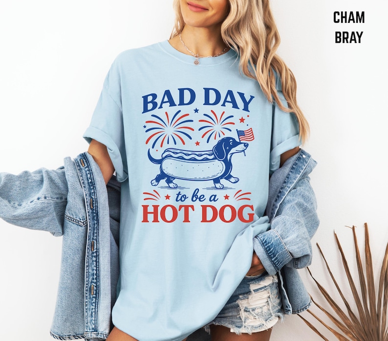 May include: Light blue t-shirt with a graphic of a dachshund hot dog, fireworks, and the text "BAD DAY to be a HOT DOG" in red and blue. The shirt is a casual style.