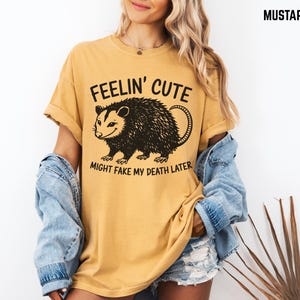 Może przedstawiać: Musztardowa koszulka z napisem "FEELIN' CUTE MIGHT FAKE MY DEATH LATER" i czarną grafiką oposa. Koszulka ma kr&oacute;tkie rękawy i luźny kr&oacute;j, odpowiednia do noszenia na co dzień.