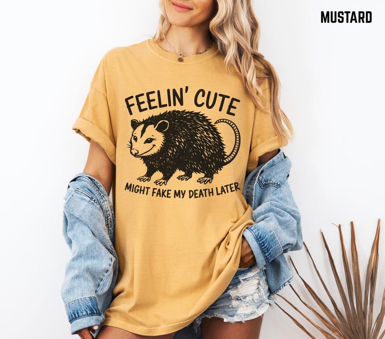 Może przedstawiać: Musztardowa koszulka z napisem "FEELIN' CUTE MIGHT FAKE MY DEATH LATER" i czarną grafiką oposa. Koszulka ma kr&oacute;tkie rękawy i luźny kr&oacute;j, odpowiednia do noszenia na co dzień.