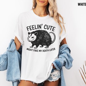Może przedstawiać: Biała koszulka z czarno-białą grafiką oposa i napisem "FEELIN' CUTE MIGHT FAKE MY DEATH LATER". Swobodny, uniseksowy styl.