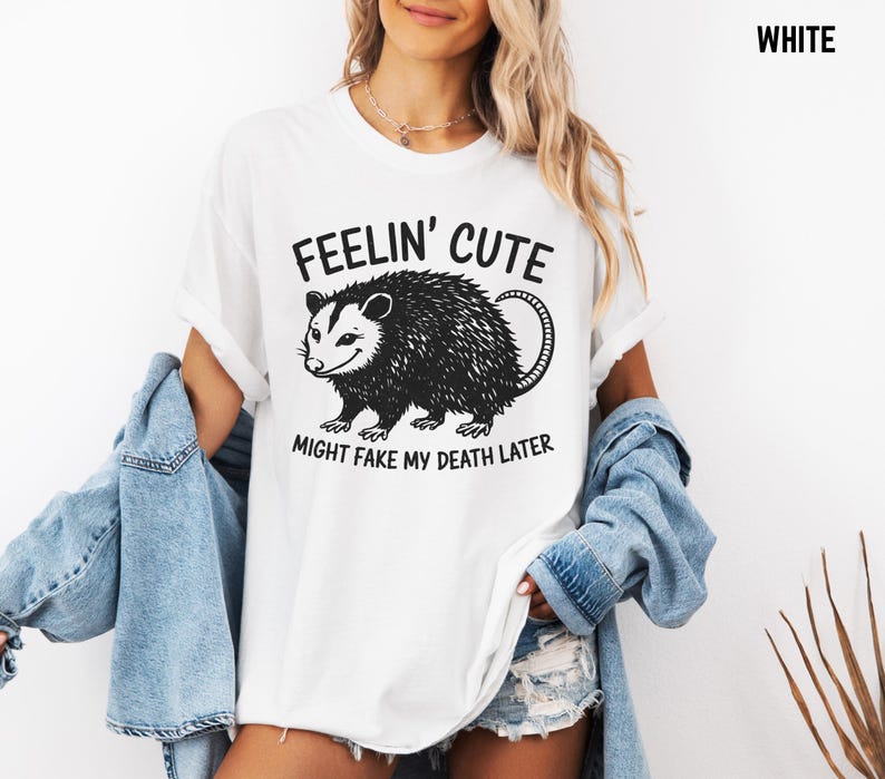 Może przedstawiać: Biała koszulka z czarno-białą grafiką oposa i napisem "FEELIN' CUTE MIGHT FAKE MY DEATH LATER". Swobodny, uniseksowy styl.