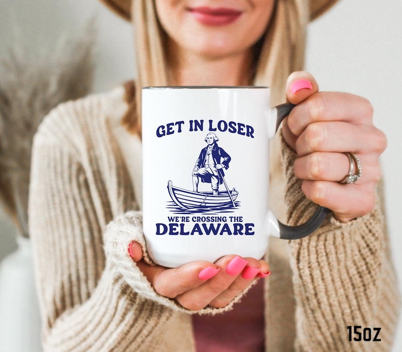 May include: White ceramic mug with a black handle and rim, featuring the text "GET IN LOSER" above an illustration of a man in a boat, with the text "WE'RE CROSSING THE DELAWARE" below. The mug is labeled "15oz".